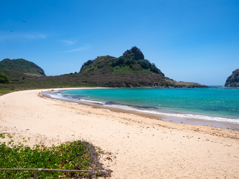 A long curved beach with teal water and white sand at Praia do Sueste on the south coast of Noronha.