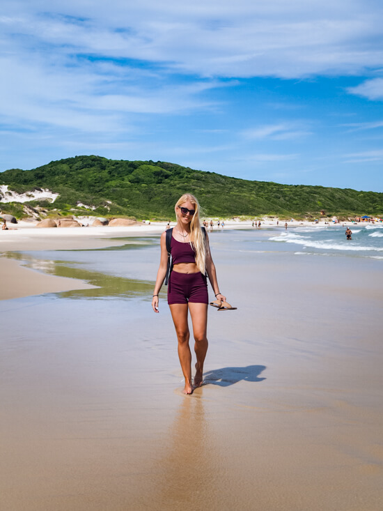 Me walking barefoot on a wide sandy beach with green hills in the background in Florianopolis.
