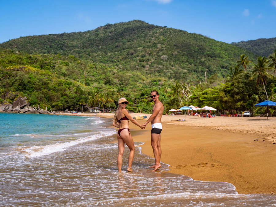 My husband and I walking along a beach with golden sand, turquoise water and green mountains in the background at Ilhabela.
