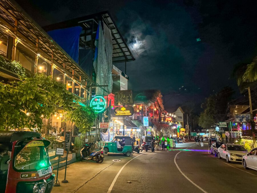 The main road of Ella lined with tuk-tuks, cafes and restaurants decorated with neon signs during the night.