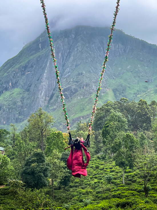 A woman wearing a red dress sitting on the giant Ella Swing decorated with flowers and overlooking the towering Ella Rock.