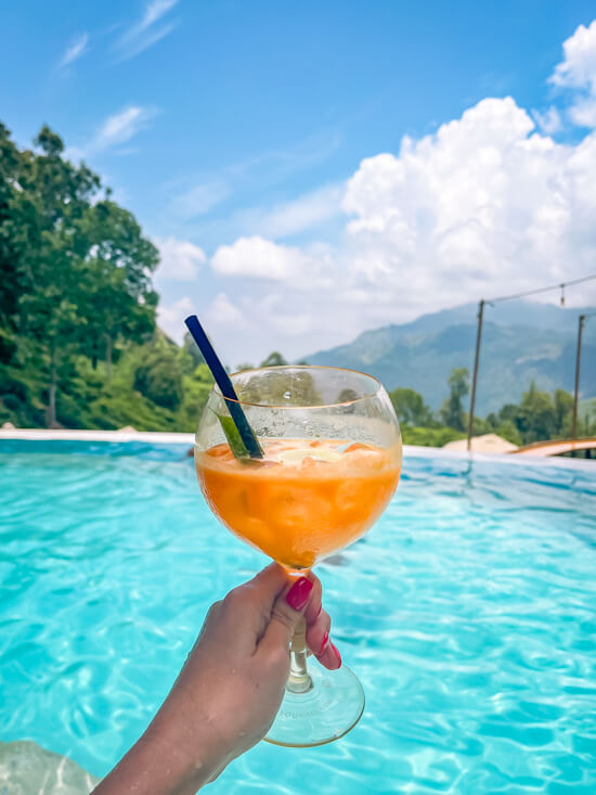 My hand holding a glass with orange cocktail in it and a pool with bright blue water in the background.