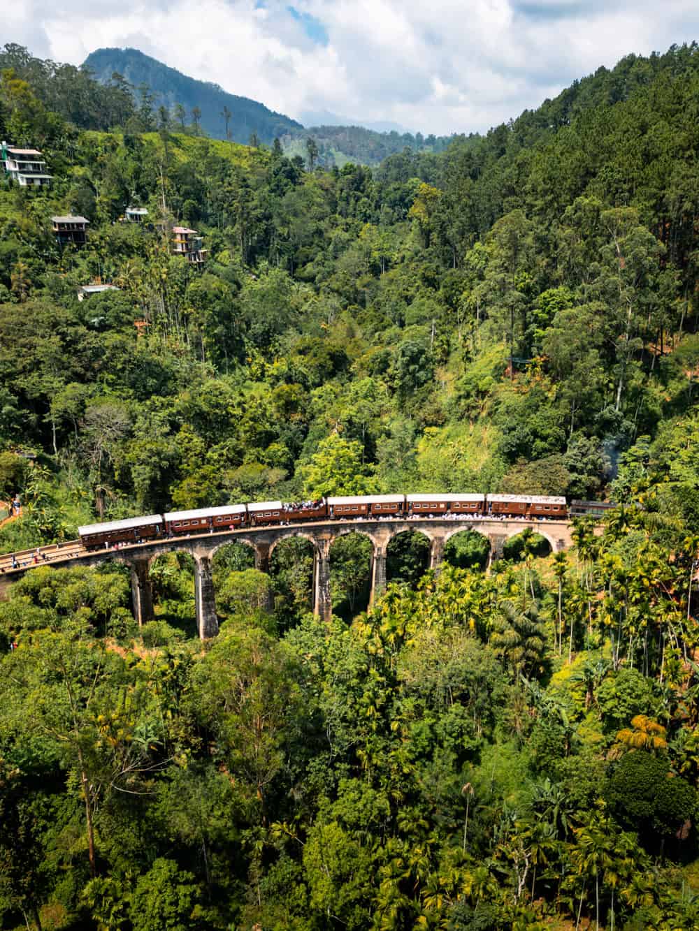 A drone photo of a train crossing the Nine Arch Bridge, surrounded by steep green hills and thick tropical vegetation.