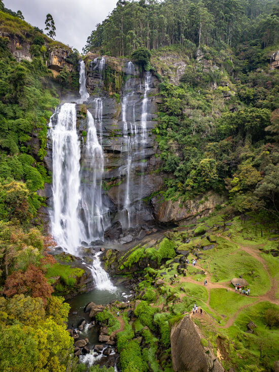 An aerial drone photo of Bomburu Ella Waterfall cascading down a black cliff surrounded by lush vegetation.