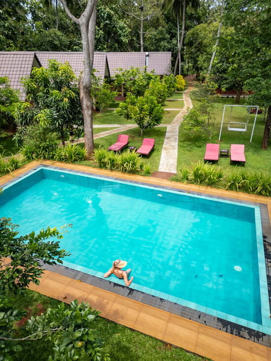 Me relaxing in a bright blue pool surrounded by a green garden and sun chairs at our hotel in Sigiriya.