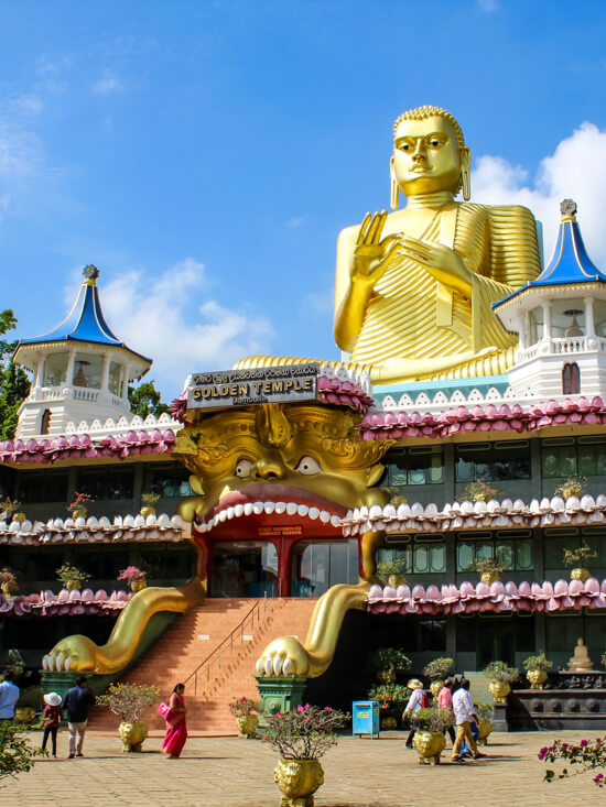 Dambulla Cave Temple adorned with colorful decorations, flowers and golden ornaments. On the roof, there's a giant golden sitting Buddha statue.