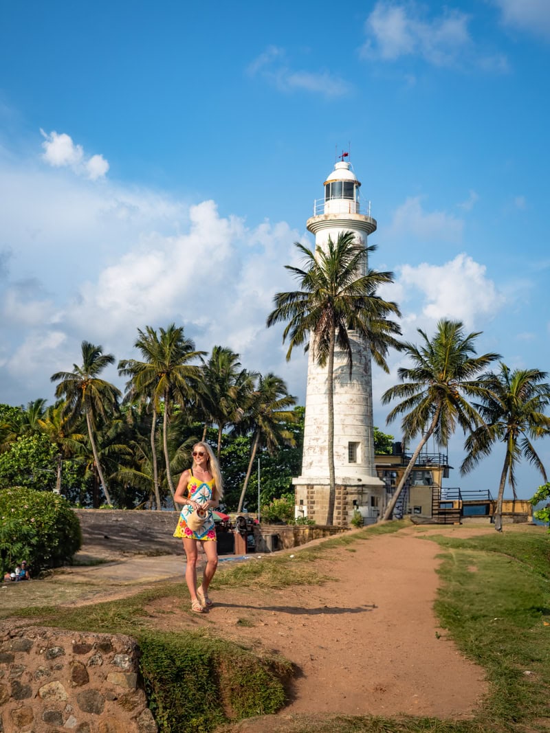 Me standing on a path along Galle Fort walls with a white lighthouse and palm trees in the background.