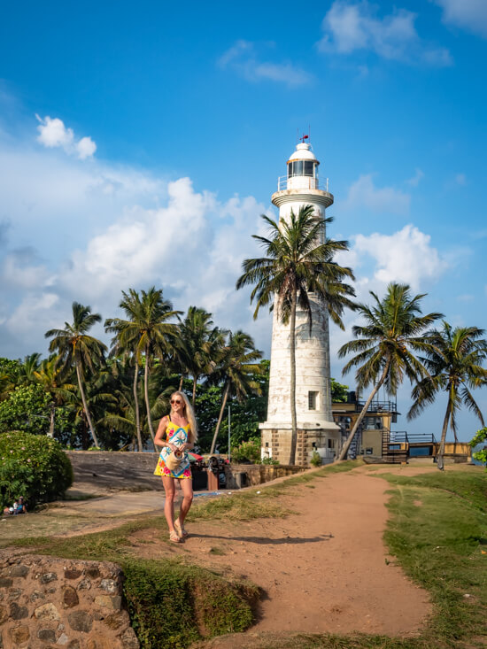 Me standing on a path along Galle Fort walls with a white lighthouse and palm trees in the background.