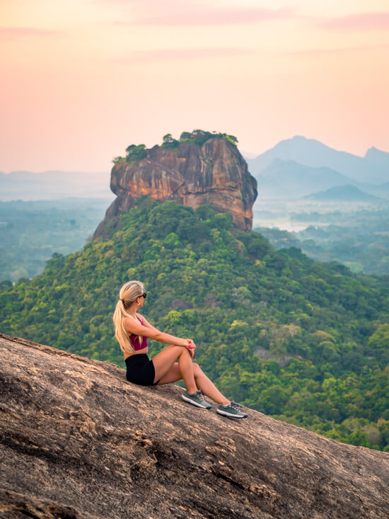 Me sitting on the rocky slope of Pidurangala Rock during sunrise with Sigiriya Rock in the background.