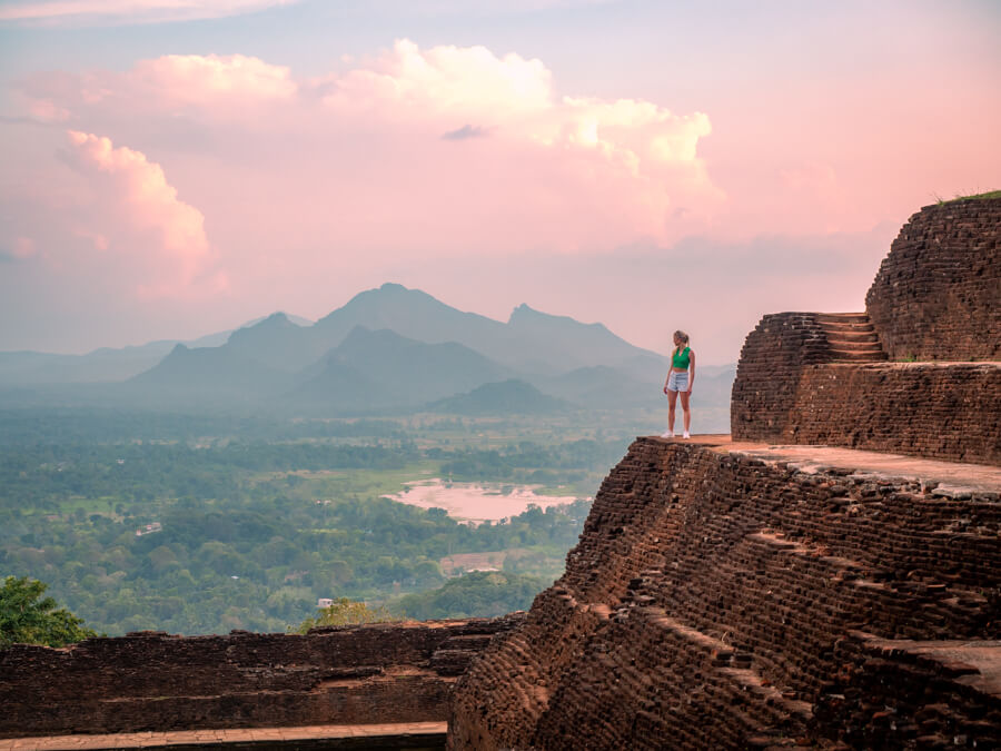 Me standing on the edge of the Sigiriya rock fortress with green mountains and pink sky in the background.