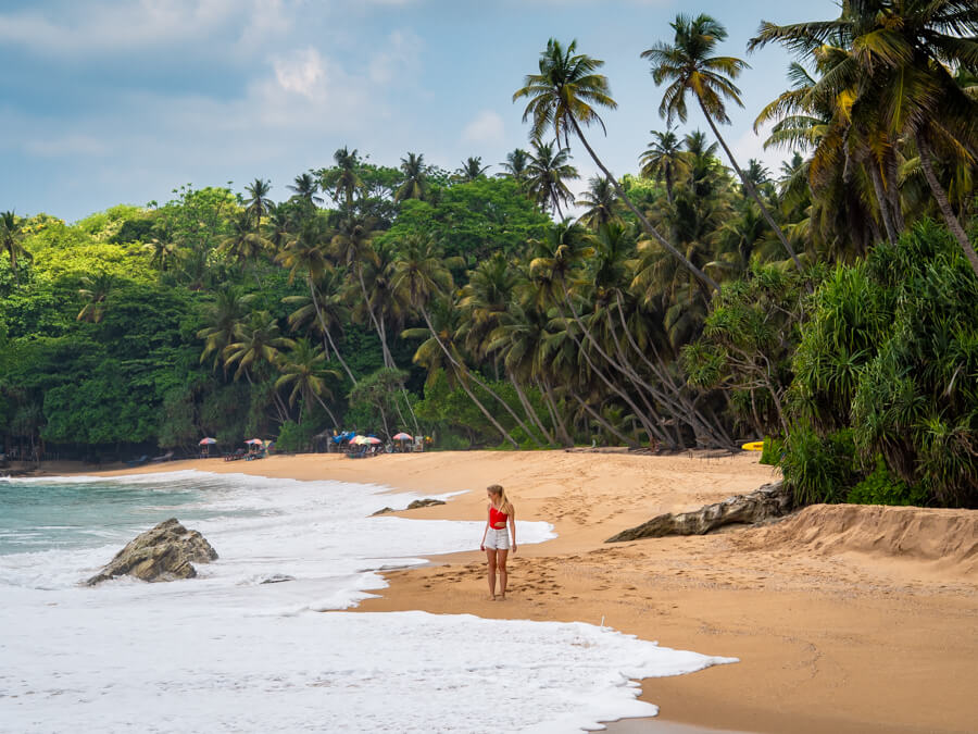 Me standing on the orange sand of Silent Beach, surrounded by white foamy waves and green palm trees leaning over the sand.