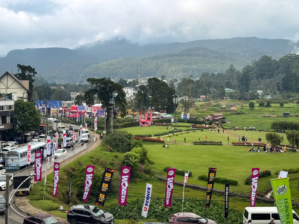 People hanging out at a park, heavy traffic on a road lined with colorful flags, and mountains covered with forest in the background in Nuwara Eliya during New Year celebrations.