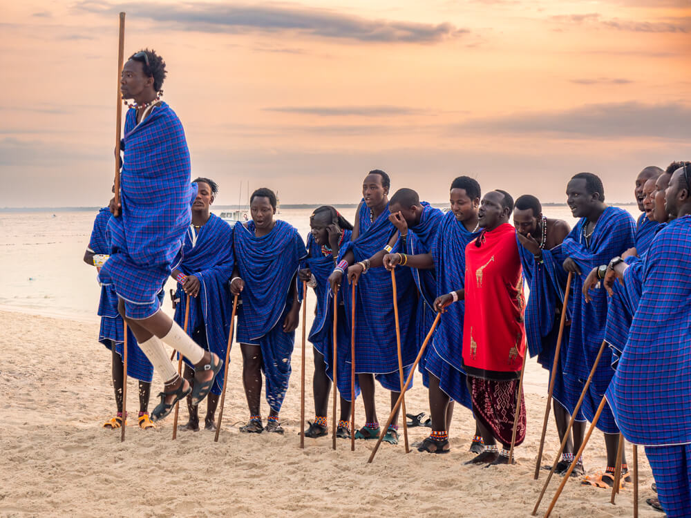 A group of Maasai men wearing blue robes and performing a traditional jumping dance on Kendwa Beach during sunset.