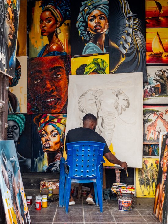 An artist sitting in his shop and painting an elephant on the canvas; he is surrounded by colorful paintings depicting African women and wildlife.