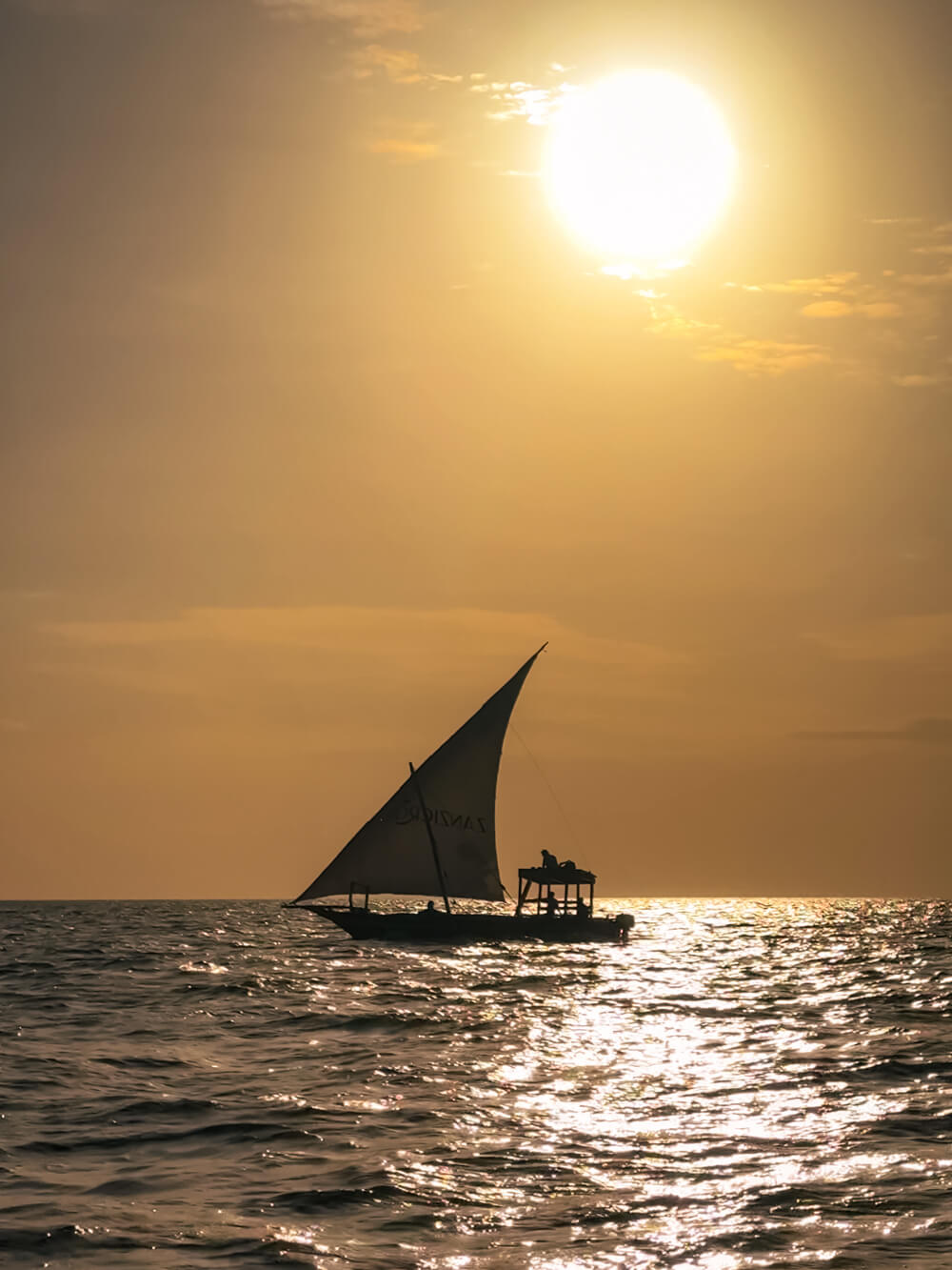 A traditional wooden dhow boat with a triangular sail sailing in the waters off the coast of Nungwi during sunset.