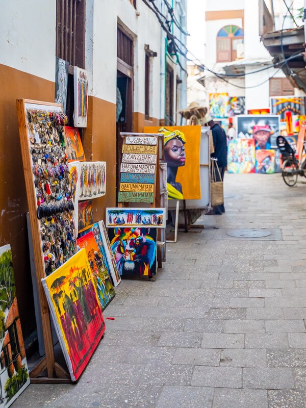 Brightly colored paintings and souvenirs displayed on a narrow street in Stone Town. 