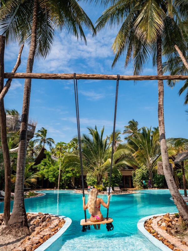 Me sitting on a swing hanging from two palm trees above a bright blue pool at The Nest Hotel in Paje.