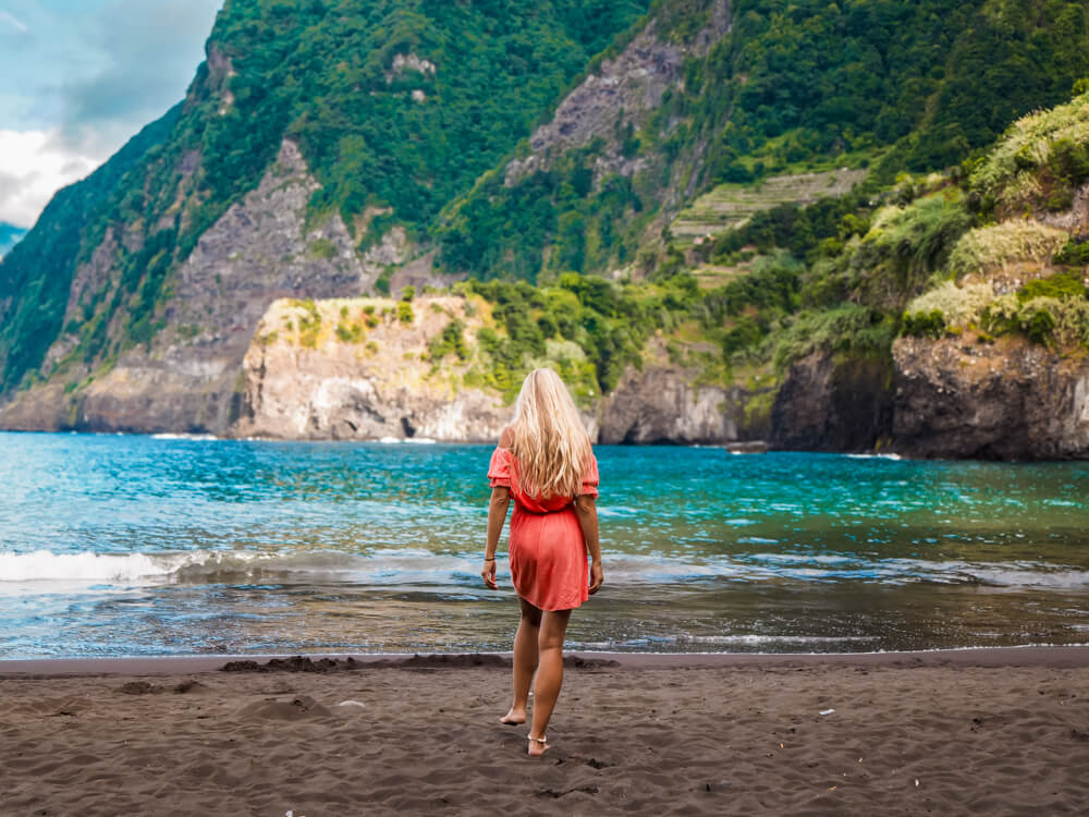 Me in a pink dress walking on the black sand of Seixal Beach in Madeira, with turquoise ocean and towering green cliffs in the background.