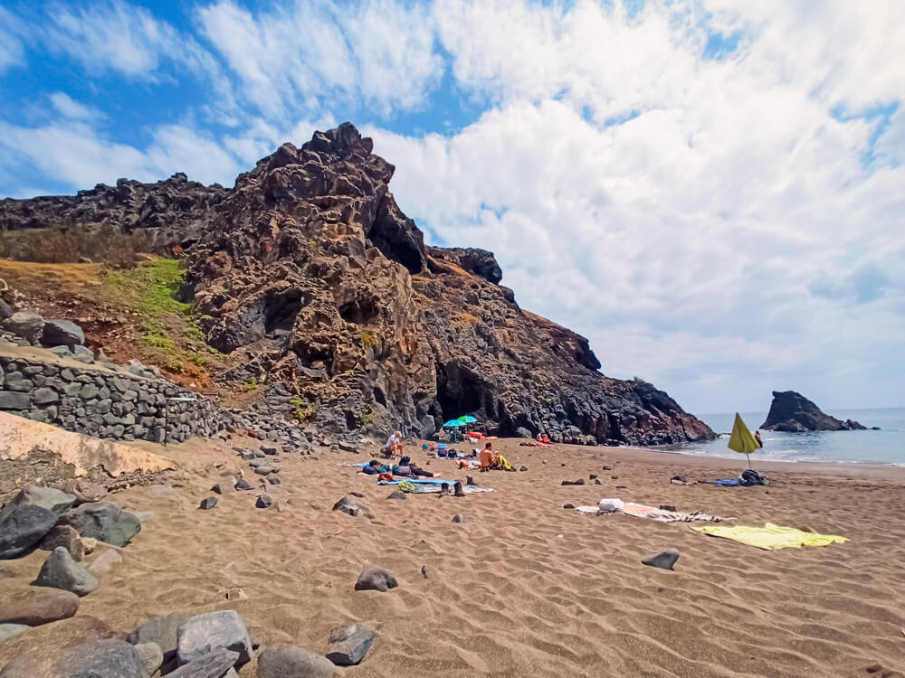Rugged volcanic cliffs, brown sand and sunbathers relaxing on towels at the secluded Prainha Beach, one of the best beaches in Madeira.