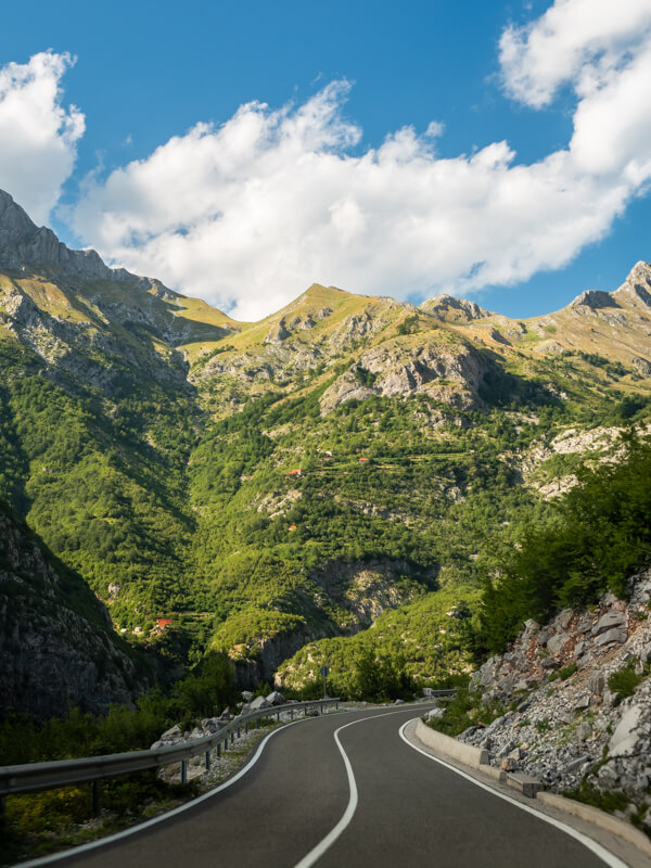 A paved road winding through the Kelmend Valley, surrounded by green mountains with jagged peaks in northern Albania.