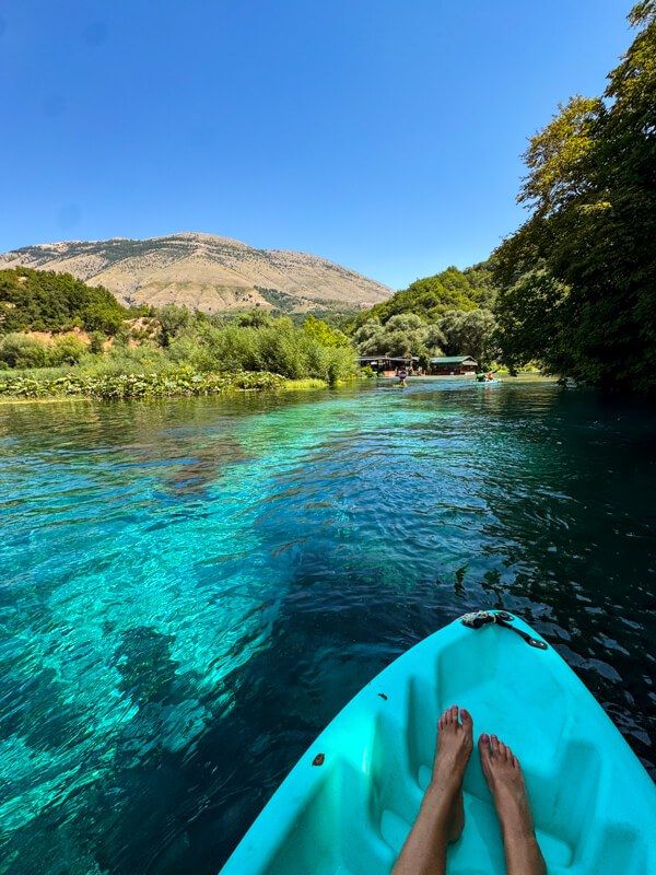Our blue kayak gliding over the crystalline turquoise waters of the Blue Eye spring with lush vegetation along its shores and a mountain in the background.