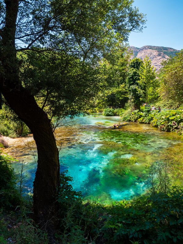 The Blue Eye spring near Saranda, Albania, with its colorful natural pool in vibrant shades of turquoise, blue and green, surrounded by trees on a sunny day.