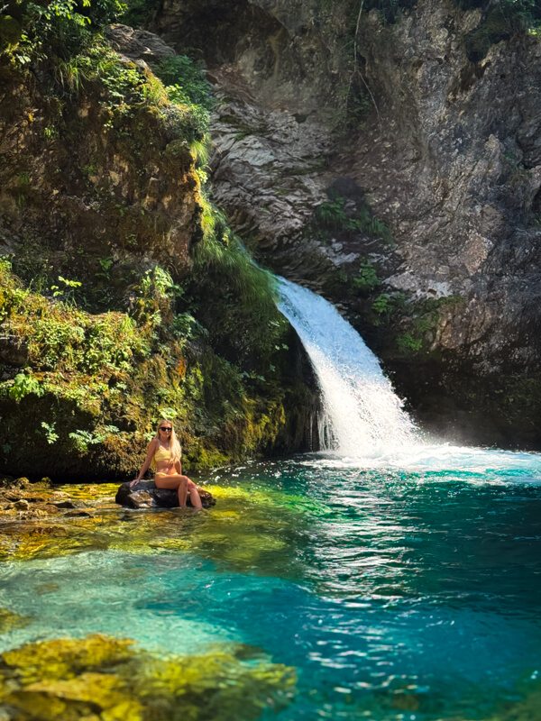 Me sitting on a rock at the Blue Eye of Theth and dangling my feet in the natural pool full of turquoise water, with a small waterfall cascading down a rock face next to me.