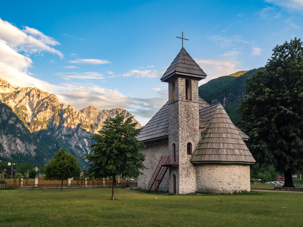 The small stone Church of Theth with a wooden shingled roof and bell tower, set against a backdrop of the Albanian Alps around Theth at golden hour.