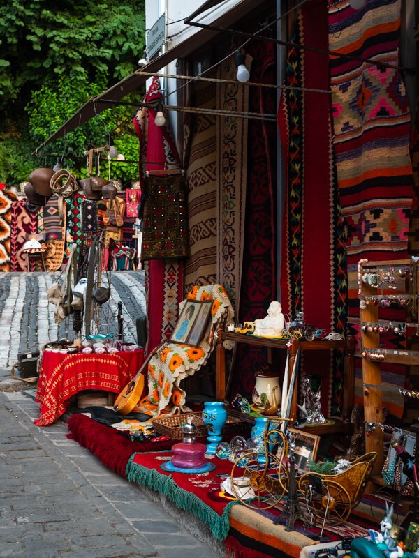 Colorful storefront of a souvenir shop in the old bazaar of Gjirokastra, displaying traditional handwoven kilim rugs, embroidered bags, vintage instruments and antique trinkets on a cobblestone street.