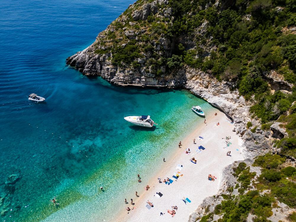 Aerial view of Grama Bay in Albania, a secluded white pebble beach surrounded by rocky cliffs with sunbathers relaxing on the shore and speedboats anchored in the vivid turquoise water.