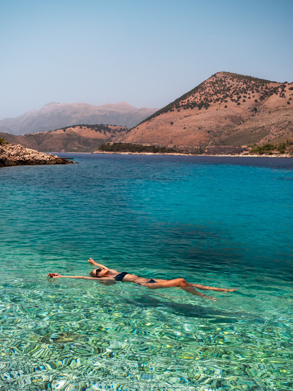 Me floating on my back in pristine turquoise water at Heart Beach in the Albanian Riviera, with arid brown hills in the background.