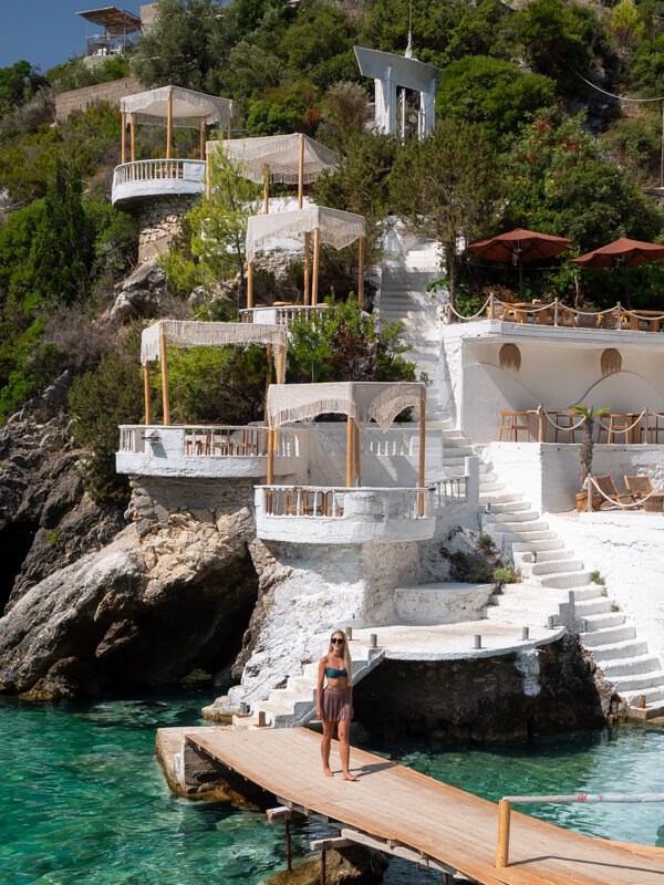 Me standing on a wooden bridge in front of the whitewashed terraces and cabanas of Kilimani Retreat, a cliffside beach club in Albania.