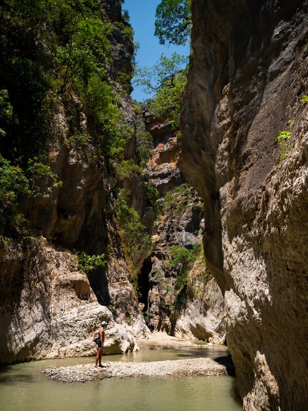My husband standing on a rocky riverbed between sheer limestone cliffs inside the narrow Langarica Canyon, with green vegetation and clear blue sky above him.
