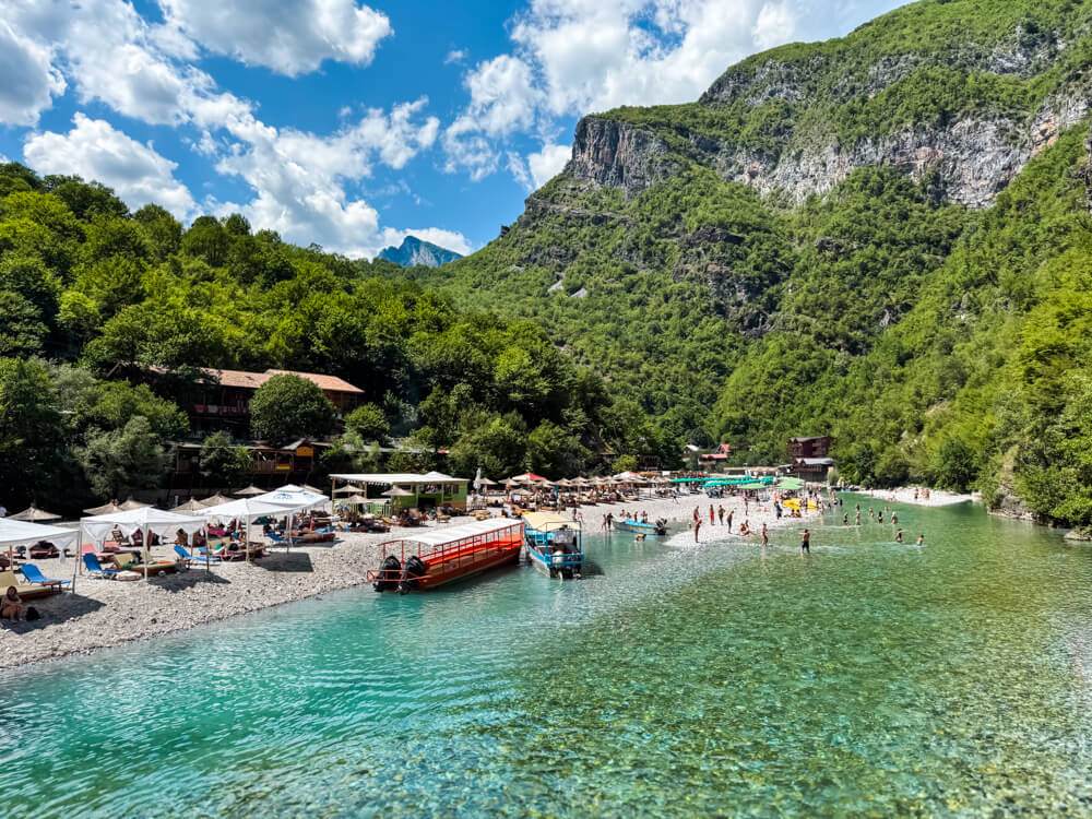 Turquoise Shala River surrounded by steep mountains covered with forest. The pebbly shores of the river are lined with sunbeds and umbrellas, and swimmers are wading in the crystal-clear water.