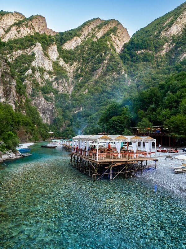 A restaurant built on stilts above the clear blue waters of Lumi i Shales river with steep jagged cliffs and lush vegetation in the background.