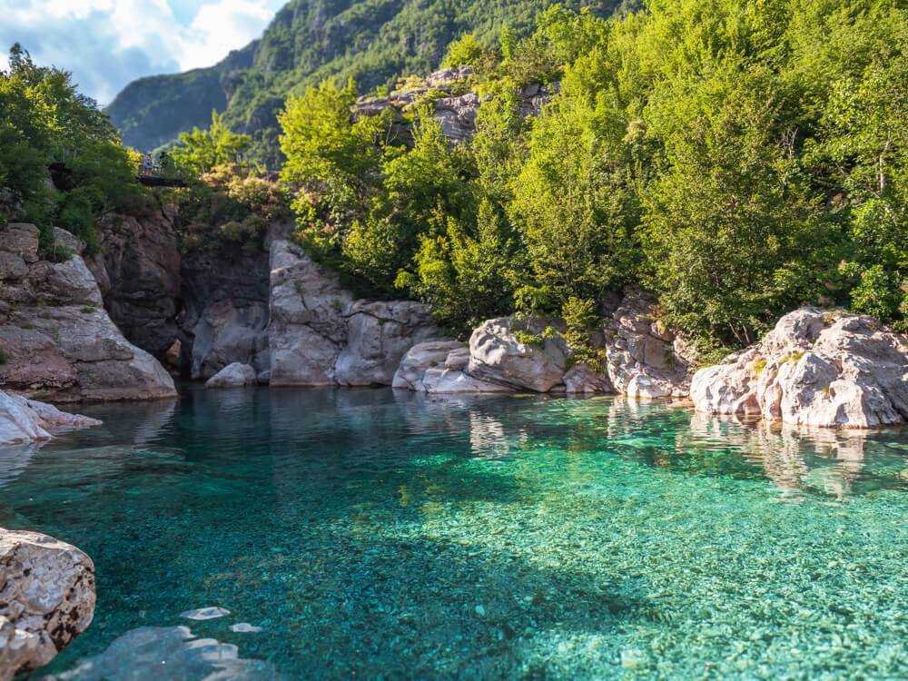 Crystal-clear turquoise natural swimming pool of Nderlysaj surrounded by smooth white boulders and green vegetation, one of the best places to visit in Theth.