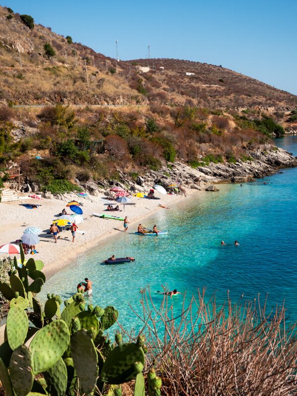 Swimmers enjoying the dazzling blue-green waters of Nemo Beach in Porto Palermo. Colorful parasols line the small white pebble beach and arid hills stretch into the distance.