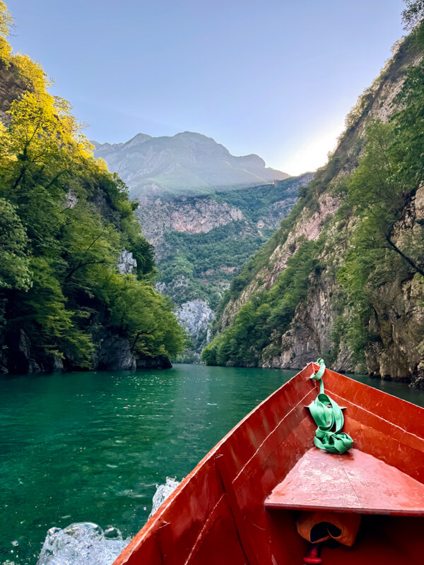 View from the bow of a red wooden boat cruising along the emerald green Shala River through a narrow canyon lined with steep cliffs and green vegetation.