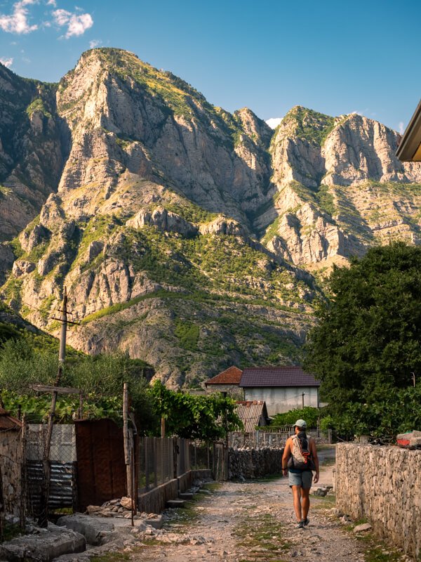 My husband walking along a dirt road in Tamara village in Albania with towering jagged mountains in the background.