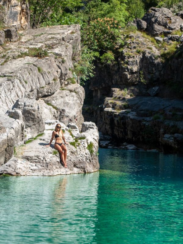 Me sitting on a rocky ledge bordering a natural river pool full of turquoise water in Tamare, Albania.
