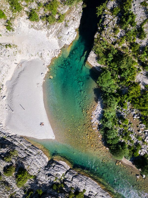 A top-down drone shot of Tamare Beach with its turquoise river pool and a small white pebble beach surrounded by green shrubs.