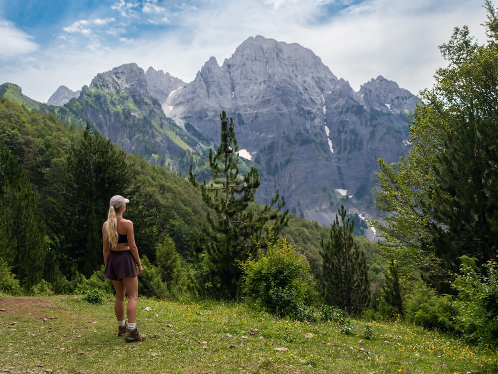 Me standing on an alpine pasture surrounded by a forest on the Theth to Valbona hike, looking up at steep rocky peaks in the Albanian Alps.