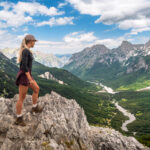 Me standing on a rocky summit at Valbona Pass in Albania, looking out over the vast green Valbona Valley with a winding river and dense forests, surrounded by the peaks of the Albanian Alps.