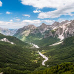 Panoramic view from Valbona Pass over the the Valbona Valley, with green forested slopes and a winding river on the valley floor, bordered by the jagged grey peaks of the Albanian Alps.