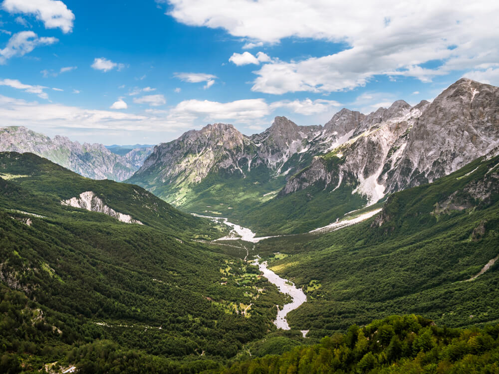 Panoramic view from Valbona Pass over the the Valbona Valley, with green forested slopes and a winding river on the valley floor, framed by the rocky grey peaks of the Albanian Alps.