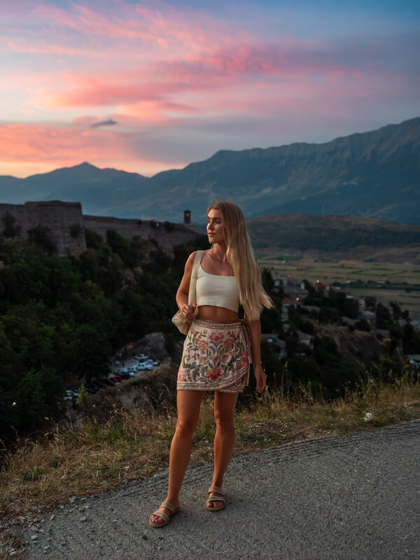 Me standing on a hill Gjirokaster, with the Castle of Gjirokastra, a mountain ridge and a sunset sky with pink and purple hues behind me.