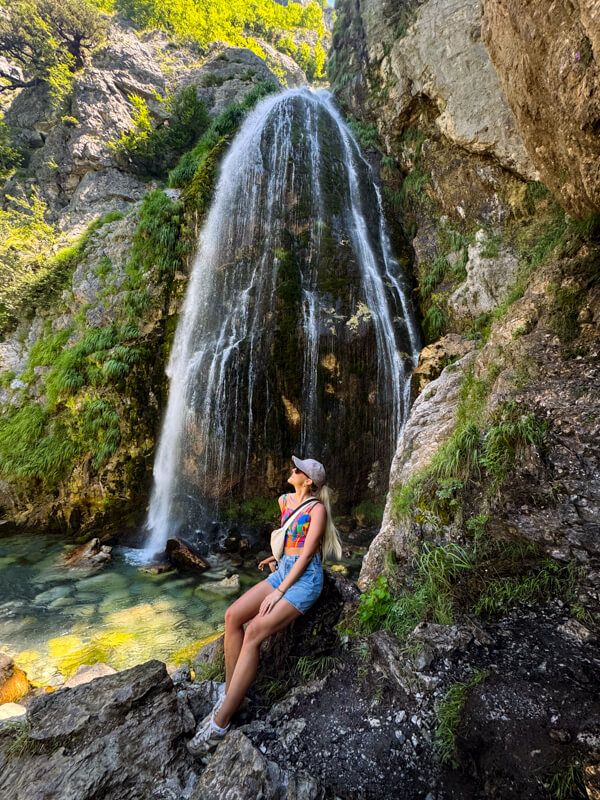 Me sitting on a rock in front of Grunas Waterfall that cascades down a rock face into a small emerald pool. 
