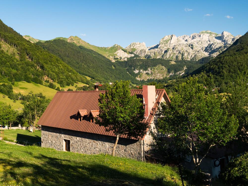 Our guesthouse in Lepushe with stone walls and a brown metal roof surrounded by trees, with green meadows, forested slopes and rocky peaks in the background.
