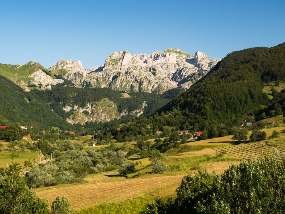 The remote mountain village of Lepushe in the Albanian Alps, set in a green valley surrounded by forested slopes and dramatic limestone peaks in the background.