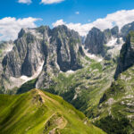 A panoramic view of rocky peaks with patches of snow and a trail along the ridge of a grassy mountain in the foreground, viewed from Maja e Vajushes, the best hike in the Albanian Alps.